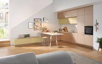 Moderne Wohnküche mit Holzfronten und gelben Oberschränken, hellem Frühstückstresen mit Hockern, daneben ein Paar beim gemeinsamen Kaffeetrinken.